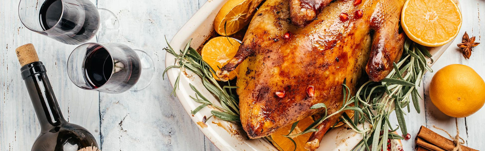 Banner mit einer Gans in Auflaufform, auf einem Holztisch und daneben zwei Weingläser